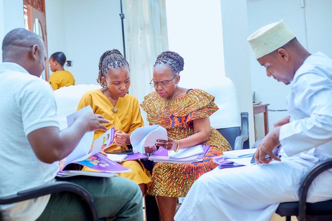 A diverse group of workshop participants actively collaborating and discussing course material around a table during an Arigatou International and GNRC Tanzania training session.
