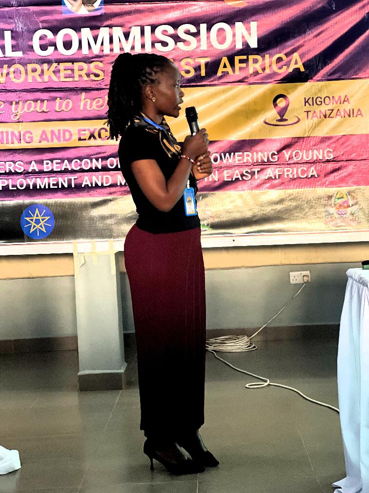 A confident keynote speaker holding a microphone and gesturing dynamically while presenting to an engaged youth audience in Kigoma.